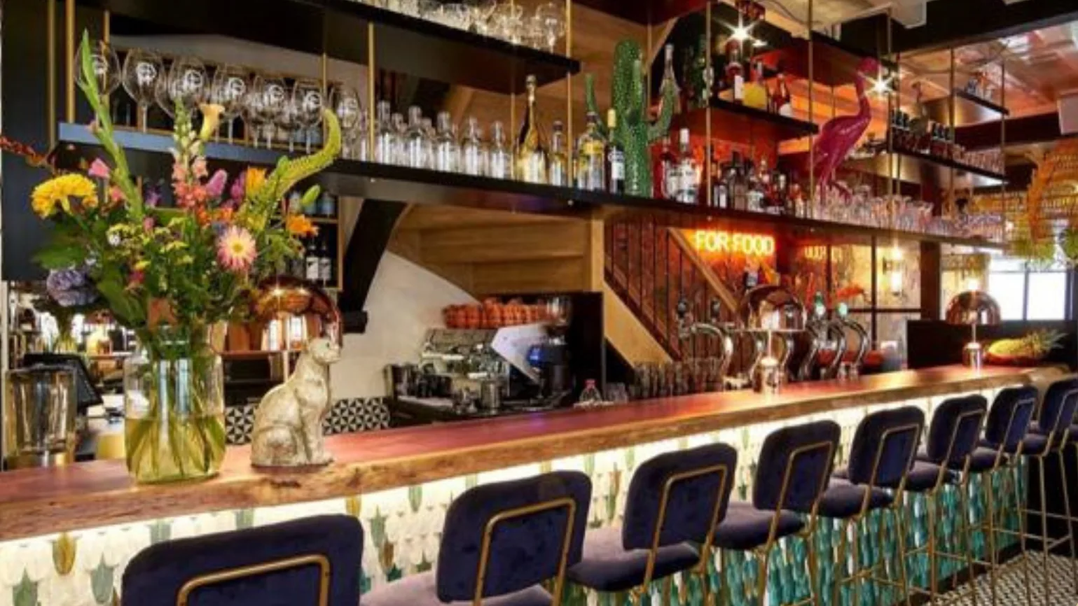 De bar van The SHOEGE, met barkrukken voor een toog van groene schubtegels en flessen op de planken